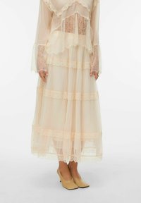 Robe beige clair transparente avec des accents en dentelle, dotée de manches longues et d'ourlets superposés et volantés. Associée à des chaussures à talons hauts beige clair.