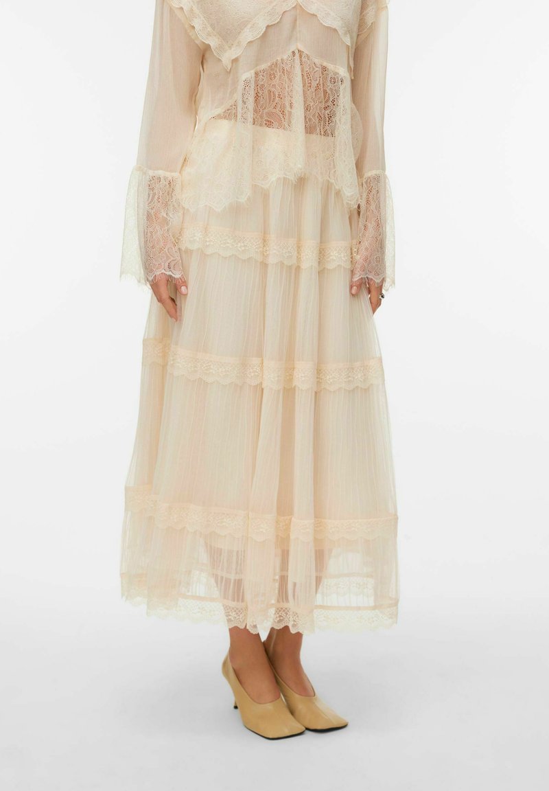Robe beige clair transparente avec des accents en dentelle, dotée de manches longues et d'ourlets superposés et volantés. Associée à des chaussures à talons hauts beige clair.