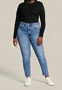 Personne portant un jean taille haute bleu clair, un haut à manches longues noir et des baskets blanches, debout devant un fond neutre.