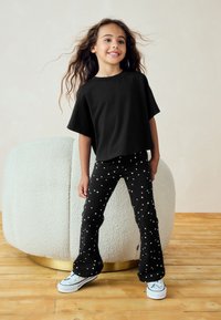 Mustad lühikeste varrukatega t-särk, mida kannavad mustad laienevad püksid, millel valged tähemustrid; valged tossud mustade detailidega. Taustal tekstureeritud ümmargune istekoht.