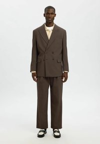 Brązowy garnitur podwójnie zapięty z szerokimi klapami, dopasowany krój, pasujące spodnie oraz kontrastujące czarne i białe buty, wyeksponowany na białym tle.