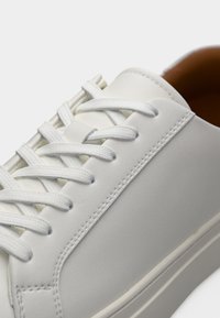 Λευκό αθλητικό παπούτσι με λείο δερμάτινο υλικό, στρογγυλεμένο σχήμα δακτύλων, πλεκτά κορδόνια και διακριτική λεπτομέρεια ραφής κατά μήκος των πλευρών και της σόλας.