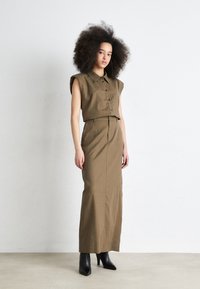 Camicia corta button-up color tan con spalle accentuate e gonna lunga abbinata. La modella indossa stivali neri con tacco alto. Lo sfondo è bianco uniforme.