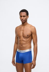 Plave bokserice s uskim krojem, mekog materijala i svijetlosmeđim pojasom s natpisom "Calvin Klein".