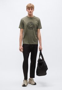 Olívazöld póló fekete, kerek grafikával, fekete nadrággal és bézs sportcipővel párosítva. Kézben tartott fekete táskászat.