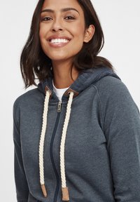 Hoodie zippé bleu marine avec une capuche texturée, des cordons de serrage crème et des accents en cuir. Tissu doux, design décontracté et fermeture éclair sur toute la longueur.