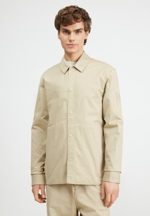 Veste légère beige avec un devant boutonné, un col plat et des manches longues. Comprend des poches latérales et une étiquette de marque discrète sur la manche.