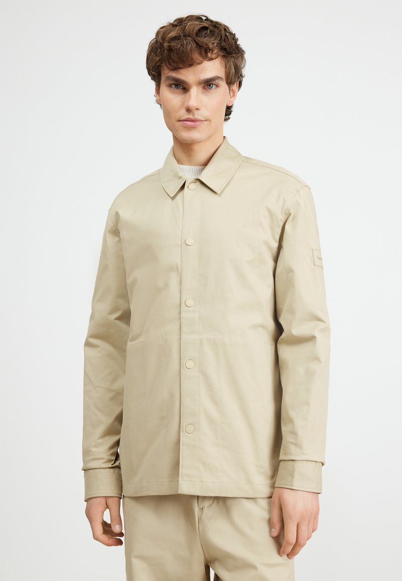 Chaqueta ligera beige con cierre de botones en el frontal, cuello plano y mangas largas. Presenta bolsillos laterales y una discreta etiqueta de la marca en la manga.