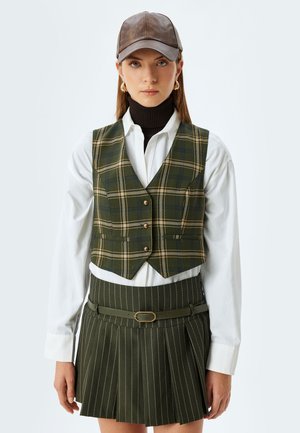 Groene geruite vest met messing knopen, gedragen over een wit overhemd, gecombineerd met een geplooide groene rok en een bruine pet. Getextureerde stof overal.