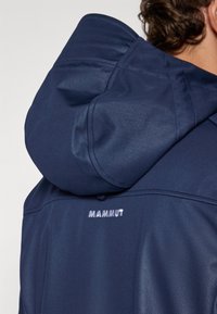 Marineblå hettejakke med glatt, elastisk stoff og brodert "Mammut"-logo bak. Designet inkluderer en strukturert hette og sømmer.