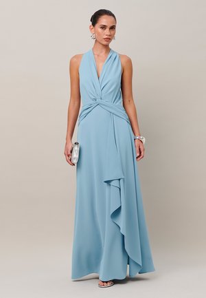 Mujer vestida con un vestido azul claro sin mangas con detalle fruncido en la cintura, sosteniendo un bolso de mano plateado y llevando joyas plateadas, de pie frente a un fondo liso.