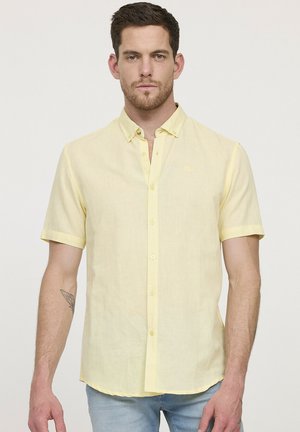 Chemise à manches courtes, de couleur jaune clair, en tissu texturé. Elle a un col et est portée avec un jean bleu clair.