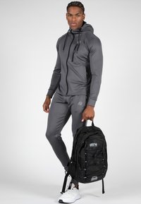 Grijze sportieve trainingspak met een rits-hoodie en taps toelopende broek. Zwarte rugzak met bungee koord details. Witte sneakers.