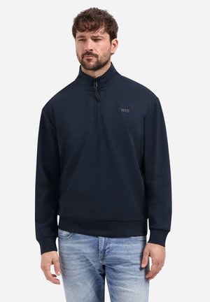 Man draagt een marineblauwe pullover met kwartrits met NXS-logo, gecombineerd met lichtblauwe verweerde jeans, staand tegen een effen achtergrond.