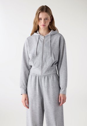 Donna con capelli lunghi che indossa una felpa grigia con zip e pantaloni della tuta abbinati con vita elasticizzata, su uno sfondo bianco uniforme.