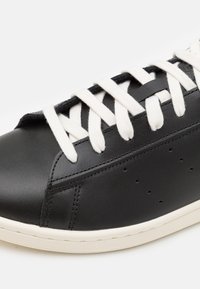 Musta nahkainen tennari, jossa valkoiset paksut nauhat, näkyvät saumat, neljä pientä ilmanottoaukkoa ja valkoinen kumipohja.