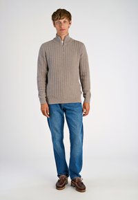 Beige gestrickter Pullover mit einem Viertelreißverschluss, gerippter Textur, langen Ärmeln, kombiniert mit hellblauen Jeans und braunen Schuhen.