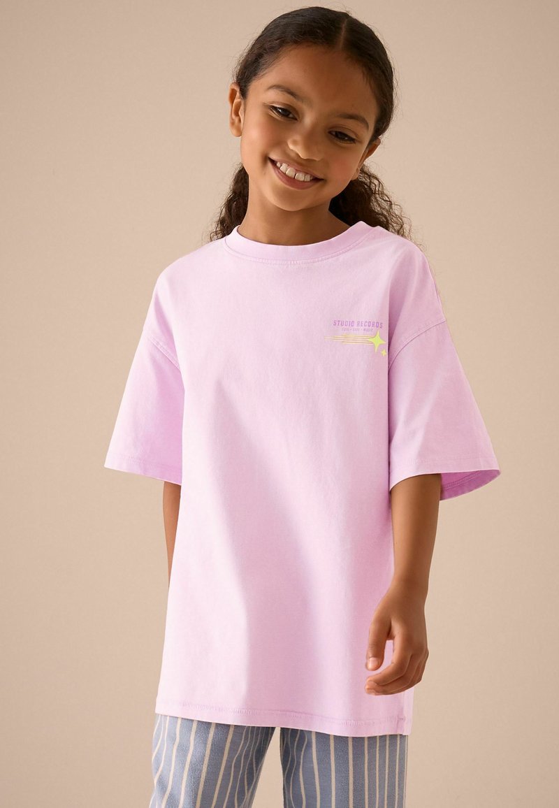 Fille souriante aux cheveux bouclés foncés porte un t-shirt rose clair ample et un pantalon rayé bleu sur un fond beige uni.