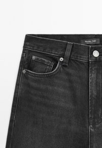 Jean en denim noir en gros plan montrant la poche avant, la passants de ceinture, les rivets argentés, le bouton, et l'étiquette Massimo Dutti à l'intérieur de la ceinture.