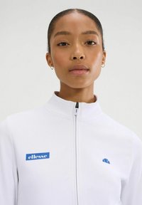 Chaqueta blanca con cremallera y cuello alto, logo azul de Ellesse en el lado izquierdo del pecho y un logo sutil en el hombro derecho. Textura suave.