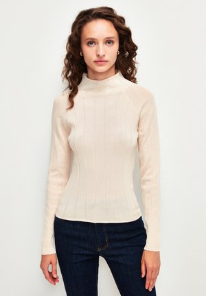 Hellbeige langärmliges, geripptes Turtleneck-Oberteil mit taillierter Silhouette, glatter Textur und subtilen vertikalen Streifen. Getragen mit dunklen Jeans.