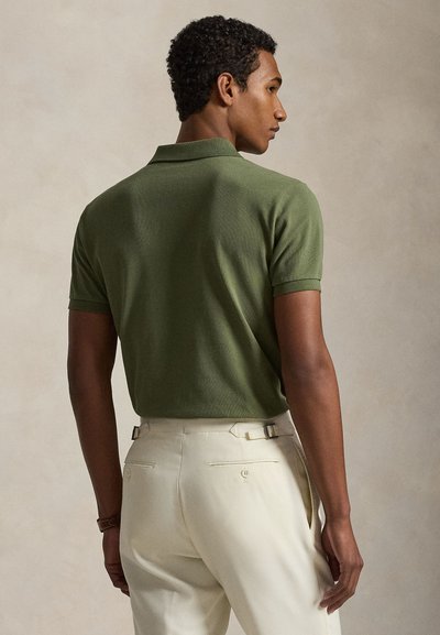 Vīrietis ar muguru vērsts, valkājot olīvu zaļu īsroku polokrekli un krēmkrāsas piegrieztas bikses neitrālā fonā.