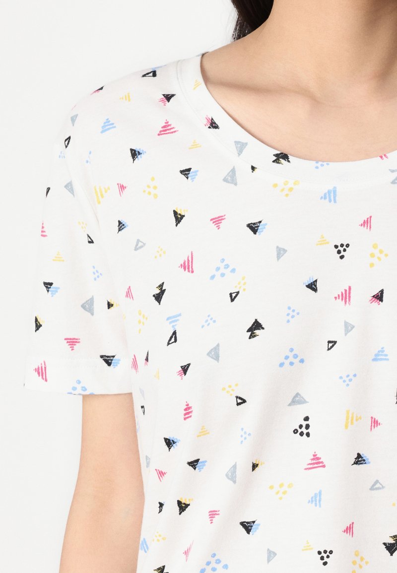 T-shirt blanc à manches courtes présentant des motifs géométriques colorés en rouge, bleu, jaune, noir et gris sur une surface de tissu lisse.