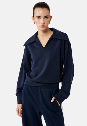 Giovane donna che indossa un top blu navy a maniche lunghe con un collo ampio e pantaloni abbinati blu navy, che posa con una mano in tasca.