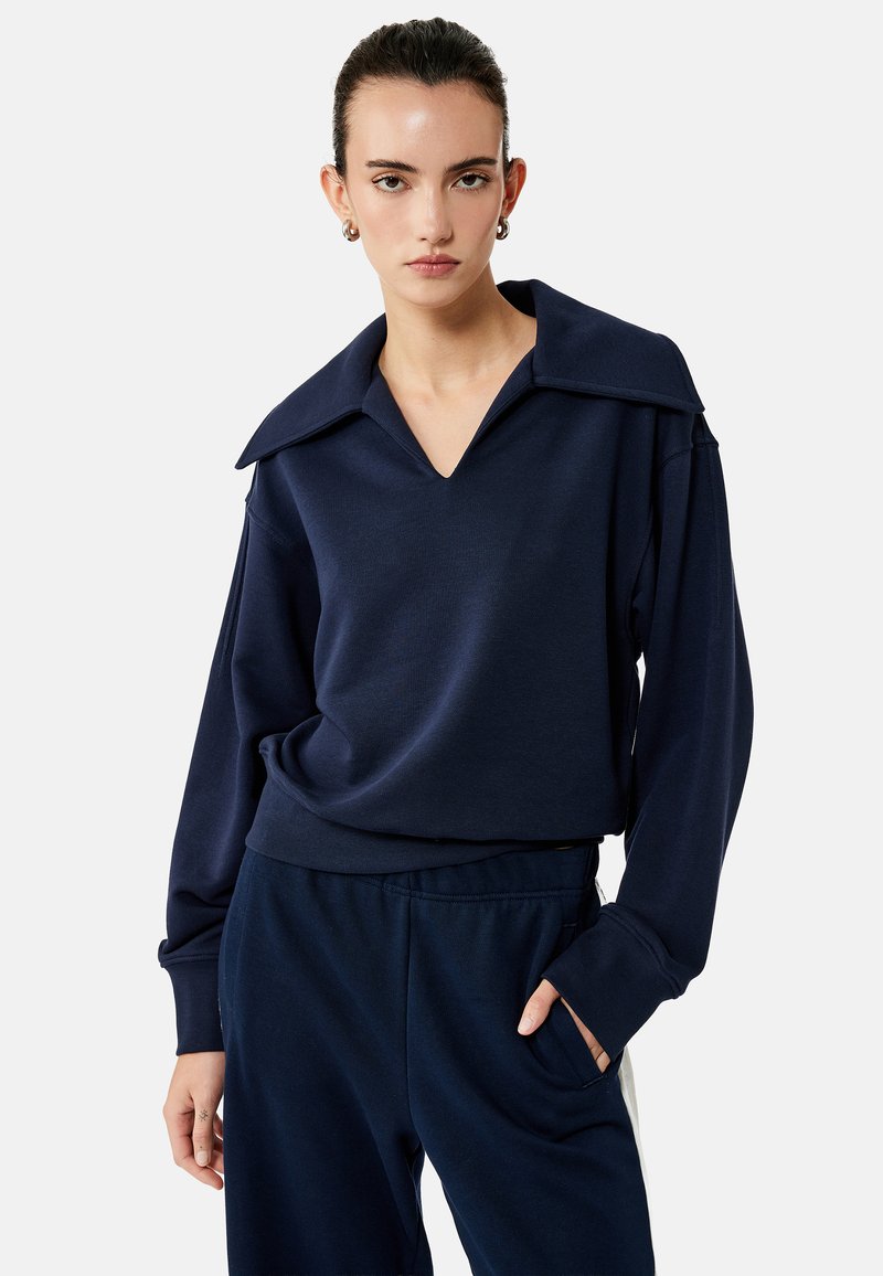 Jonge vrouw draagt een marineblauwe top met lange mouwen en een brede kraag en een bijpassende marineblauwe broek, poseert met één hand in de zak.
