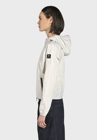 Leichte, cremefarbene Kapuzenjacke mit elastischen Bündchen und einer seitlichen Reißverschlusstasche. Mit einem Logo-Patch auf dem linken Ärmel.