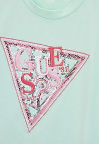 Camiseta verde menta con un gran logotipo triangular de lentejuelas rosas y plateadas que presenta las letras "GUESS" y "81" en varias fuentes y texturas.