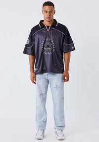 Jersey viola scuro a maniche corte con dettagli rosa e grafica centrale. Jeans azzurri con motivi di stelle ricamati. Sneakers bianche.