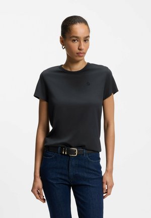 Kobieta o krótkich włosach, ubrana w czarny dopasowany T-shirt włożony w ciemnoniebieskie jeansy z czarnym paskiem, stojąca zwrócona przodem na białym tle.