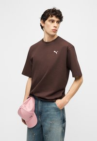 Νέος άνδρας φορώντας καφέ μπλουζάκι Puma και μπλε τζιν, κρατώντας ένα ροζ καπέλο μπέιζμπολ με το ένα χέρι, ενώ το άλλο χέρι είναι στην τσέπη του.