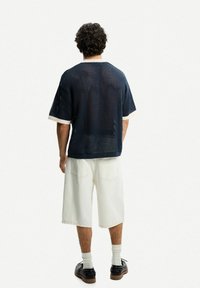 Maglietta a maniche corte in rete blu navale con accenti crema, abbinata a pantaloni bianchi e scarpe scure, vista da dietro. Fit rilassato e testurizzato.