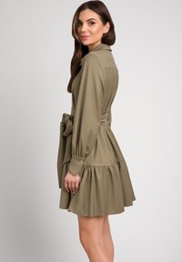Khaki sukienka koszulowa z długimi rękawami, wiązanym w pasie i warstwową spódnicą. Posiada zapięcia na guziki przy mankietach oraz kołnierz, co nadaje jej uporządkowany wygląd.