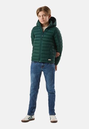 Junge steht mit den Händen hinter dem Rücken, trägt eine grüne Kapuzen-Steppjacke, blaue Jeans und weiße Turnschuhe mit grünen Streifen.