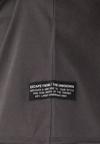 Tejido de camiseta gris oscuro con textura acanalada, que presenta una etiqueta rectangular negra con texto blanco que indica la marca y el eslogan cosida en el lateral.