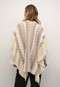 Beige og creme mønstret poncho med zigzag-design, frynser i kanten og tekstureret strik; hænger over skuldre og midt på ryggen.