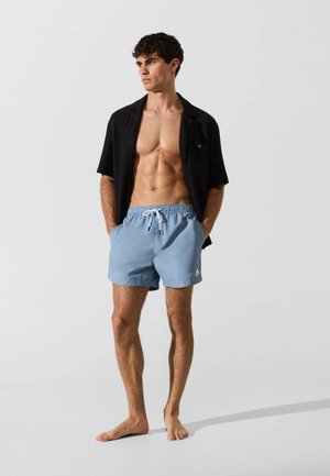 Muž, ktorý má na sebe otvorenú čiernu košeľu s krátkym rukávom a svetlomodré plavky, stojí bosý a pozerá sa do strany na jednofarebnom svetlom pozadí.