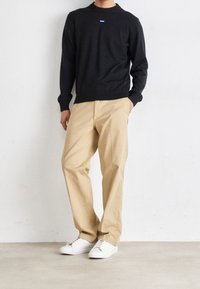Mann in schwarzem langärmeligen Pullover, beigen Hosen und weißen Sneakers, der mit einer Hand in der Tasche an einer schlichten weißen Wand steht.