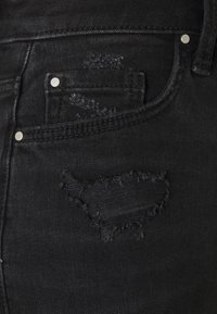 Pantalones vaqueros de mezclilla negra con una zona desgastada y bordes deshilachados, herrajes plateados y un diseño clásico de cinco bolsillos.