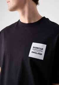 Zwarte katoenen t-shirt met een wit rechthoekig label waarop in dikke letters de tekst "BARBOUR INTERNATIONAL SOUTH SHIELDS ENGLAND" staat.