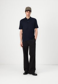 Polo shirt bleu marine à manches courtes et col, associé à un pantalon noir à jambes larges. Le modèle porte une casquette à carreaux marron et des chaussures noires.