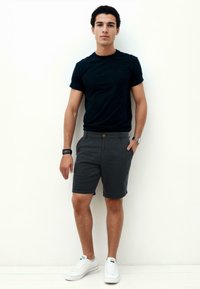 Black short-sleeve t-shirt, gray shorts with a button closure, and white sneakers, standing against a light background.