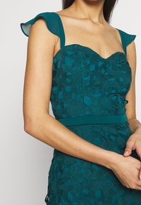 Tealfärgad klänning med blommig spetsöverlägg, hjärtformad halsringning och utsvängda ärmar. Figursydd midja och texturerad yta, med ett slätt tygband.