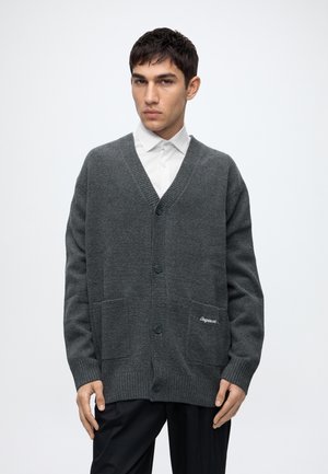 Jovem usando um cardigan cinzento escuro com botões por cima de uma camisa branca e calças pretas, em pé contra um fundo claro e simples.