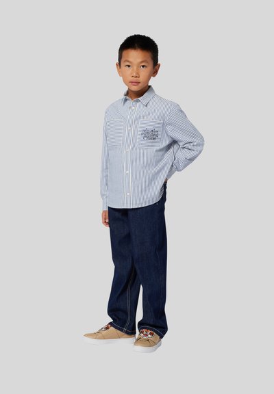 Chemise boutonnée bleu clair à rayures blanches avec deux poches plaquées, assortie à un jean bleu foncé et des baskets beiges avec des détails brodés.