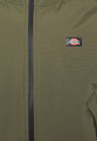 Chaqueta verde oliva con cierre de cremallera, tejido texturizado, cremallera negra y un prominente parche con el logo de Dickies. Cuenta con bolsillos laterales.