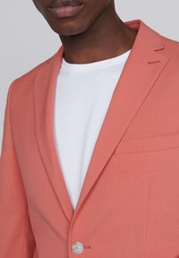 Blazer corallo realizzato in tessuto leggero con chiusura a un bottone, rever a pattina e taschino sul petto, sopra una t-shirt bianca a girocollo.